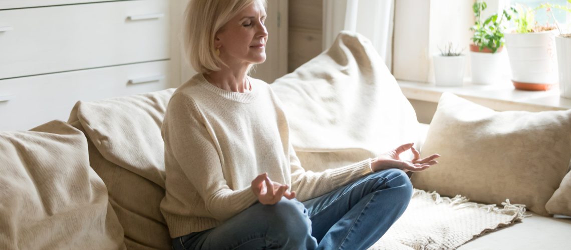 Mindful healthy mature woman practicing meditation at home, calm senior middle aged lady sitting on couch in lotus pose doing yoga for mental balance breathing air relaxing on stress free weekend